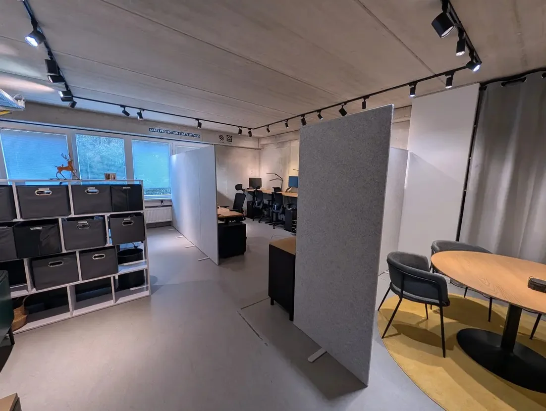 Büro von ADence in Hamburg: Arbeitsplätze mit Monitoren hinter grauen Akustikwänden, Sichtbetondecke mit Strahlern, links ein Regal mit Aufbewahrungsboxen, rechts ein runder Besprechungstisch auf gelbem Teppich